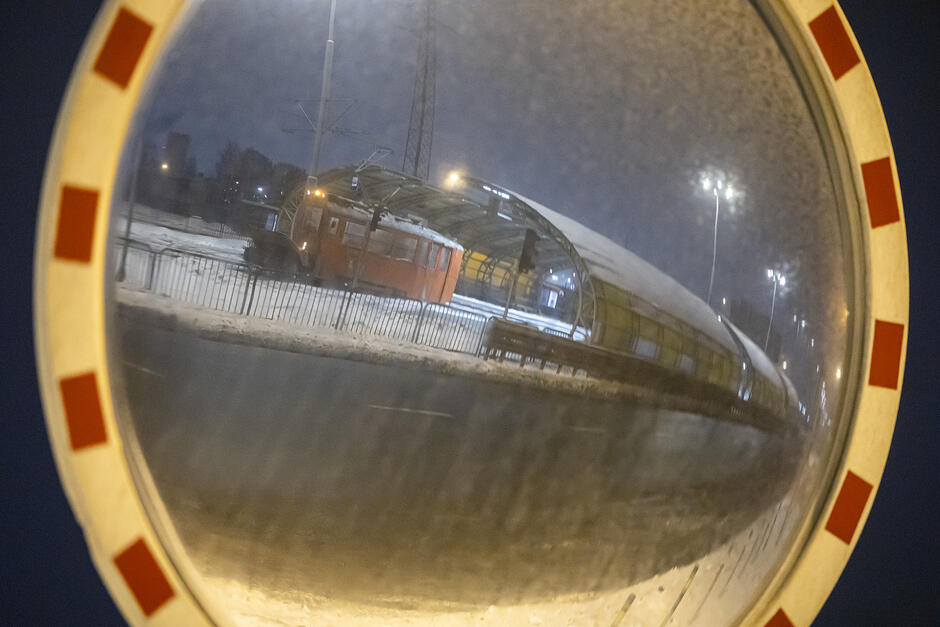 Zdjęcie przedstawia widok w lustrze drogowym, w którym odbija się nowoczesna, przeszklona wiata przystankowa lub tunel nad trasą szybkiego ruchu w zimowej scenerii. Wewnątrz zadaszonej konstrukcji widoczny jest pomarańczowy pojazd, prawdopodobnie techniczny lub tramwaj, stojący na ośnieżonym torowisku pod osłoną nocy