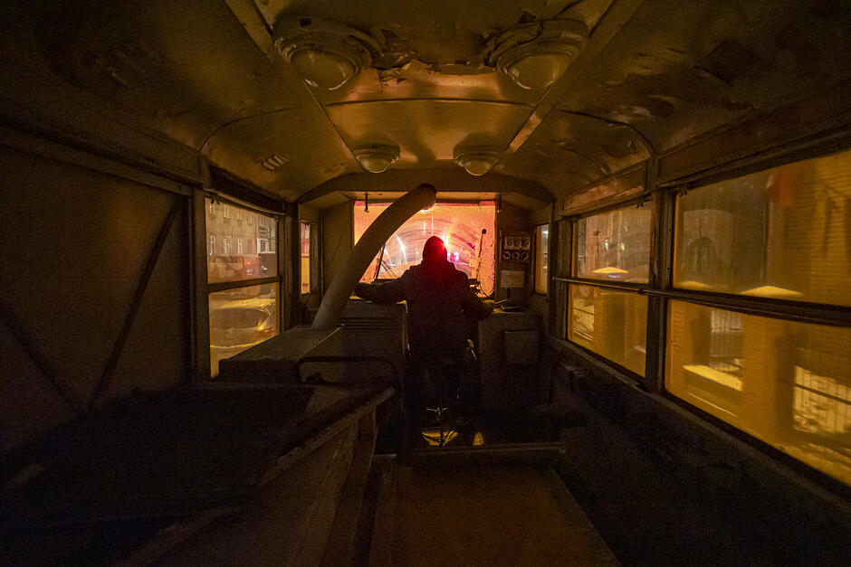 Zdjęcie przedstawia mroczne wnętrze starego tramwaju lub wagonu technicznego, w którym centralną postacią jest motorniczy siedzący tyłem do widza. Przez przednią szybę widać rozświetlony czerwonym światłem tunel, tworzący kontrast z ciepłym, żółtym oświetleniem sufitowym pojazdu.