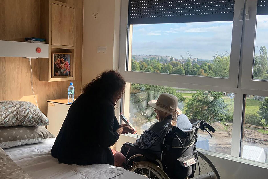 Dwie kobiety siedzą przed oknem, jedna stara siedzi na wózku inwalidzkim, druga młodsza na łóżku obok i mierzy starszej ciśnienie.