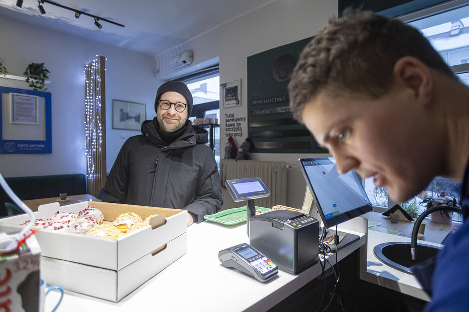 Zdjęcie przedstawia klienta kupującego pączki w kawiarni, przy ladzie wyposażonej w terminal płatniczy i system sprzedażowy. W tle widać jasne, nowocześnie urządzone wnętrze z tablicą informującą o serwowaniu kawy „ze szczyptą tolerancji”.

