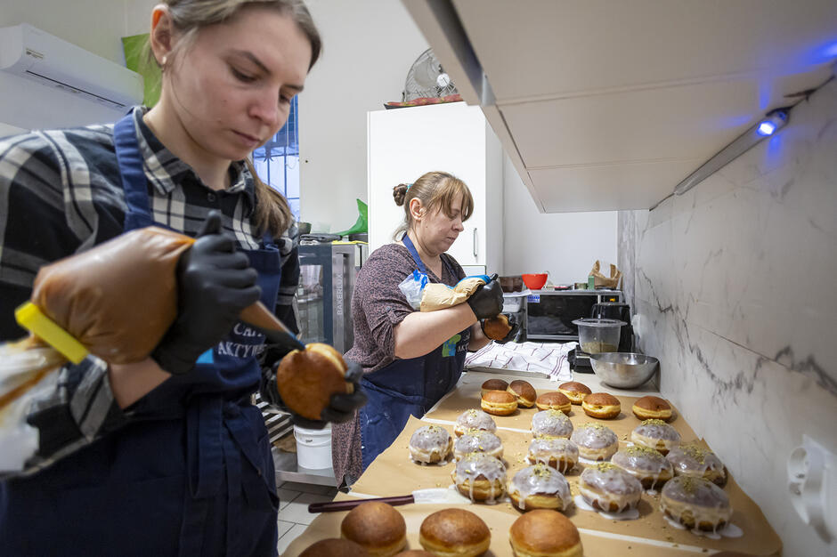 Zdjęcie przedstawia dwie pracownice kawiarni „Caffe Aktywni” podczas przygotowywania pączków w profesjonalnej kuchni. Kobiety w skupieniu nadziewają i dekorują lukrem świeże wypieki, które są apetycznie ułożone na blacie.