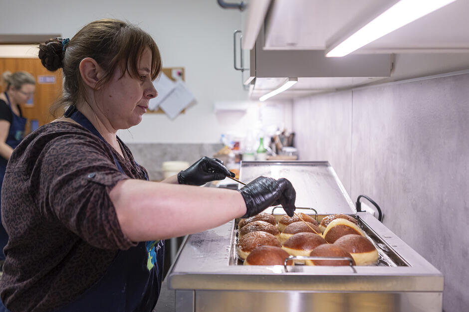 Zdjęcie przedstawia kobietę w czarnych rękawiczkach, która za pomocą specjalnych patyczków obraca pączki smażące się w głębokim tłuszczu. Proces odbywa się w profesjonalnej kuchni przy użyciu dużej, gastronomicznej frytownicy.