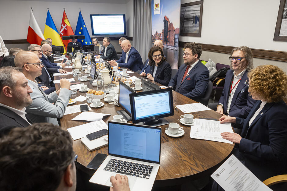 Kilkanaście osób siedzi wokół owalnego stołu na naradzie.