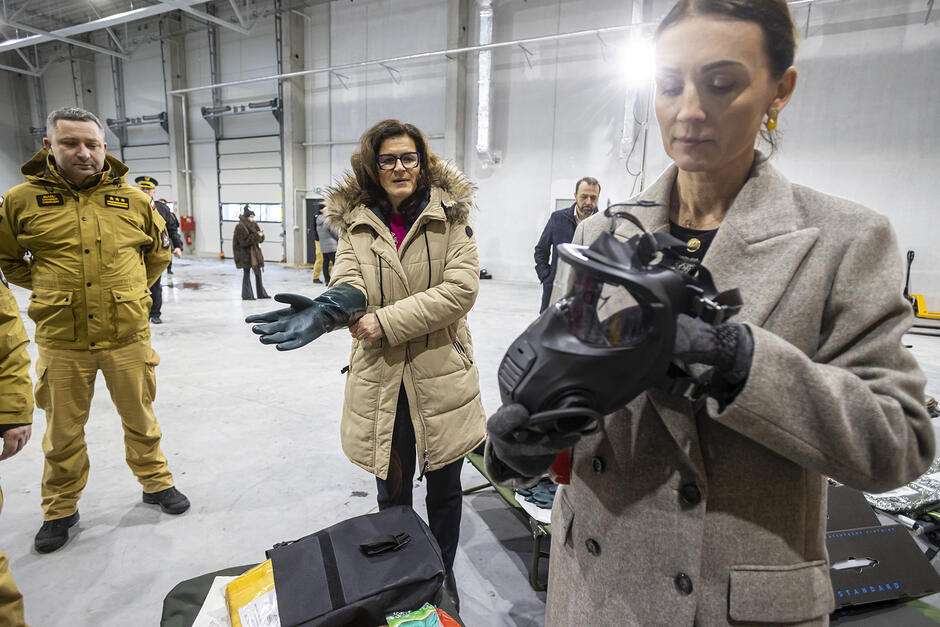 Dwie kobiety w magazynie, jedna sprawdza maskę przeciwgazową, druga wkłada rękawicę ochronną. 