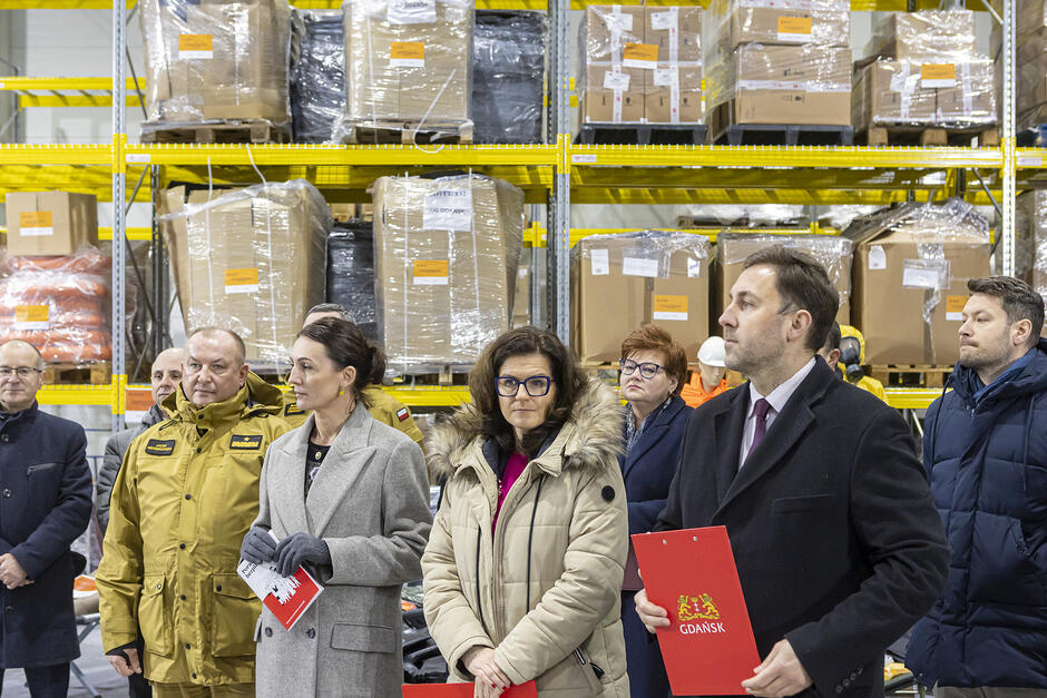 Dwie kobiety i dwóch mężczyzn, w tym jeden w mundurze strażackim, stoją w rzędzie podczas briefingu prasowego w magazynie.