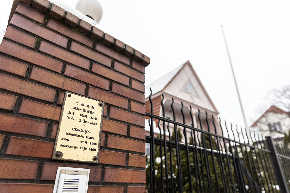 Tabliczka metalowa koloru złotego na ceglanym murku z informacją o pracy konsulatu chińskiego, w tle budynek za płotem.