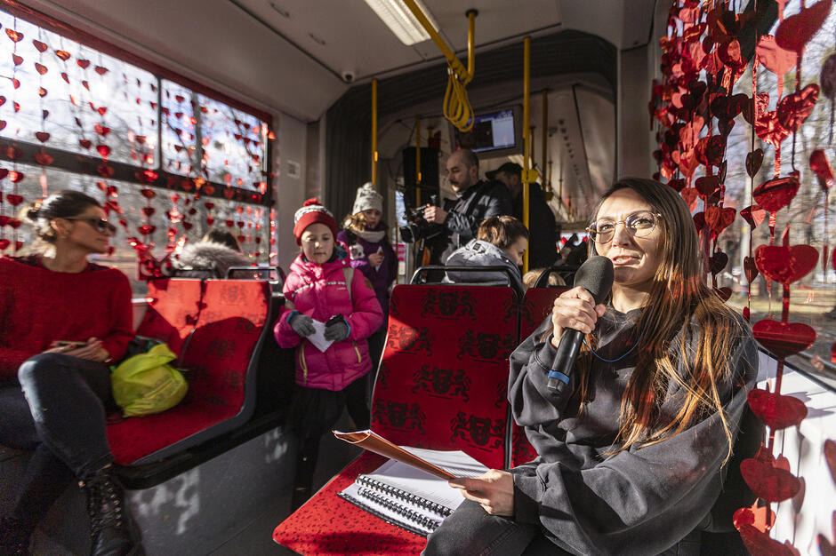 Zdjęcie przedstawia uśmiechniętą kobietę trzymającą mikrofon i teczkę z notatkami we wnętrzu tramwaju udekorowanego girlandami z czerwonych serc. W tle widać innych pasażerów oraz siedzenia z charakterystycznym wzorem gdańskich lwów, co wskazuje na specjalne wydarzenie walentynkowe w komunikacji miejskiej.