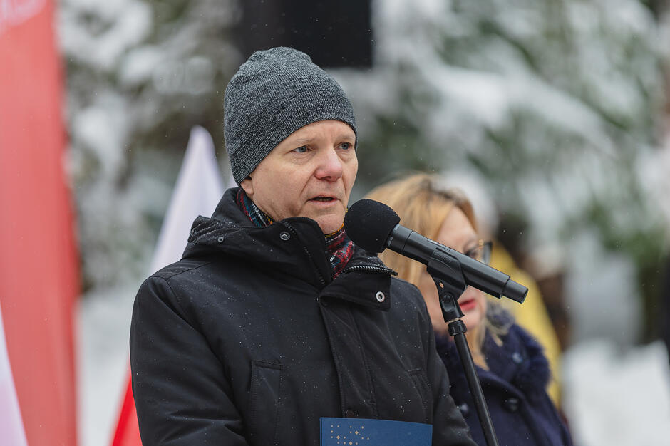 Na zdjęciu widać mężczyznę w ciemnej zimowej kurtce i szarej czapce, który przemawia do mikrofonu podczas wydarzenia na zewnątrz. W tle widoczne są biało-czerwone barwy, a cała scena rozgrywa się w zimowej, lekko zaśnieżonej scenerii.