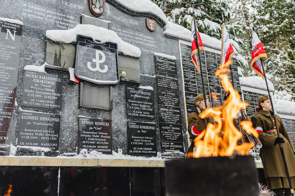 Na zdjęciu widać uroczyste obchody Narodowego Dnia Pamięci Żołnierzy Armii Krajowej, odbywające się przy Kwaterze Akowskiej na Cmentarzu Łostowickim w Gdańsku. Przed pamiątkową ścianą z nazwiskami dowódców płonie symboliczny znicz, a obok czuwają rekonstruktorzy w historycznych mundurach z polskimi flagami.