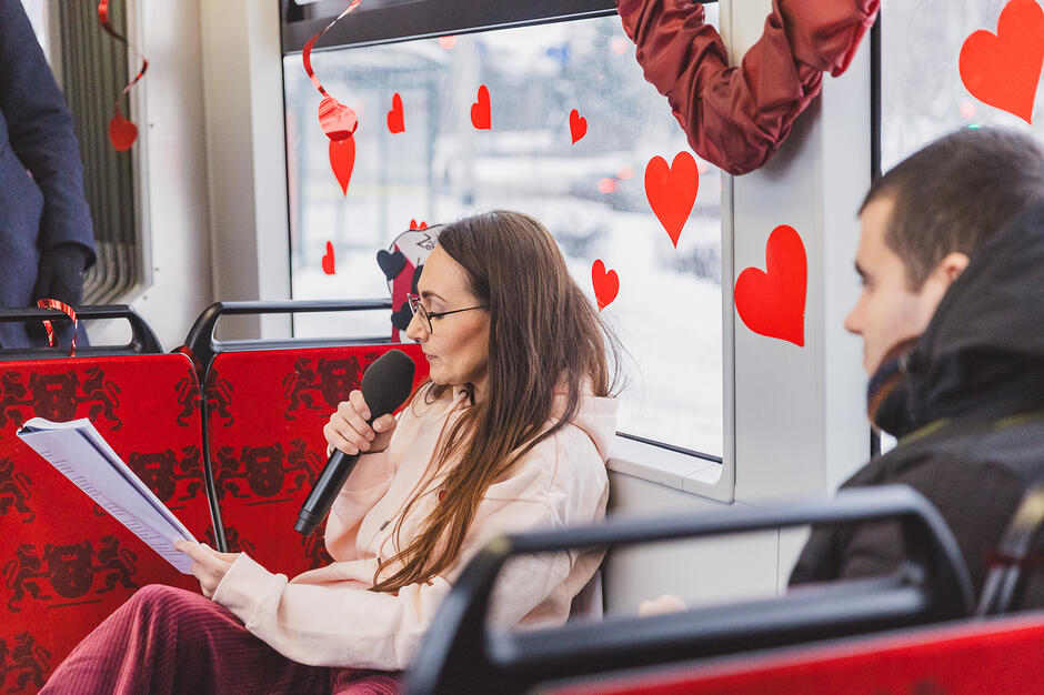 Na zdjęciu widać kobietę z mikrofonem siedzącą w tramwaju udekorowanym czerwonymi sercami, która czyta tekst z trzymanych w ręku kartek. Obok niej siedzi pasażer, a cała sceneria sugeruje specjalne wydarzenie zorganizowane z okazji walentynek.