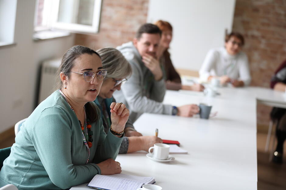 Zdjęcie przedstawia grupę osób uczestniczących w spotkaniu lub warsztatach przy długim, białym stole. Na pierwszym planie widać kobietę w okularach i zielonym swetrze, która z zaangażowaniem coś tłumaczy, podczas gdy reszta uczestników uważnie jej słucha.
