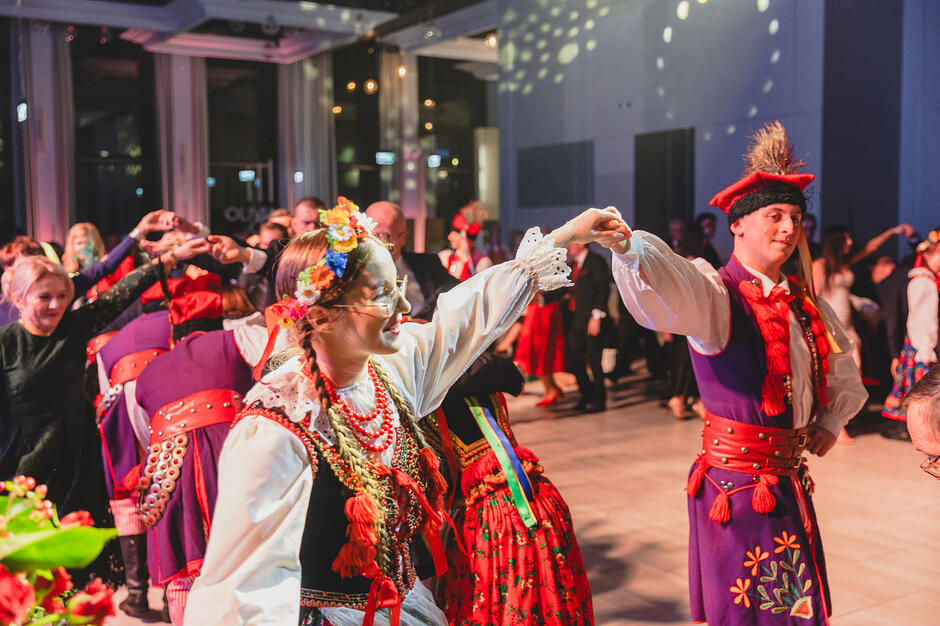 Zdjęcie przedstawia parę tańczącą w tradycyjnych strojach krakowskich podczas uroczystego balu. Kobieta ma na sobie bogato zdobiony gorset, czerwone korale i wianek z kwiatów, natomiast mężczyzna nosi charakterystyczną rogatywkę z pawim piórem.
