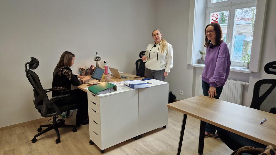 Trzy panie. Jedna siedzi przed komputerem przy biurku, dwie stoją i patrzą w obiektyw
