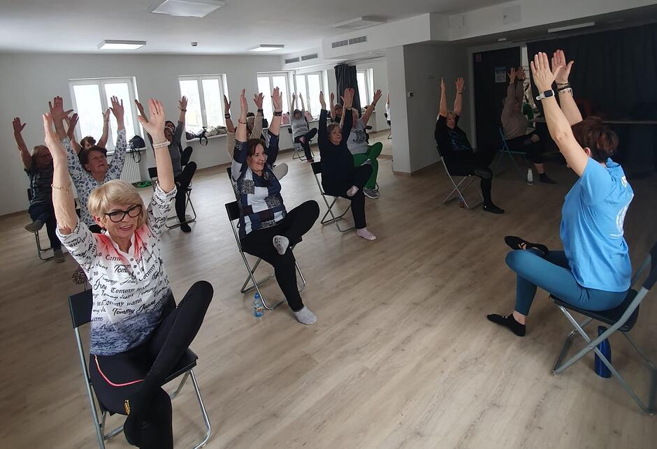 Starsze osoby siedzące na krzesłach w sali gimnastycznej, podnoszące w górę ręce