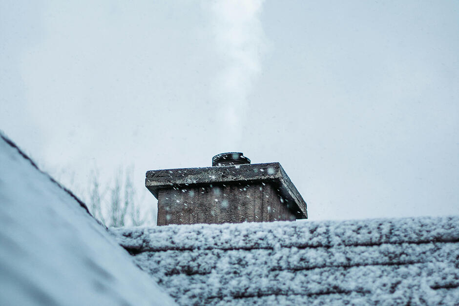 Zaśnieżony spadzisty dach domu z kominem, z którego leci dym.