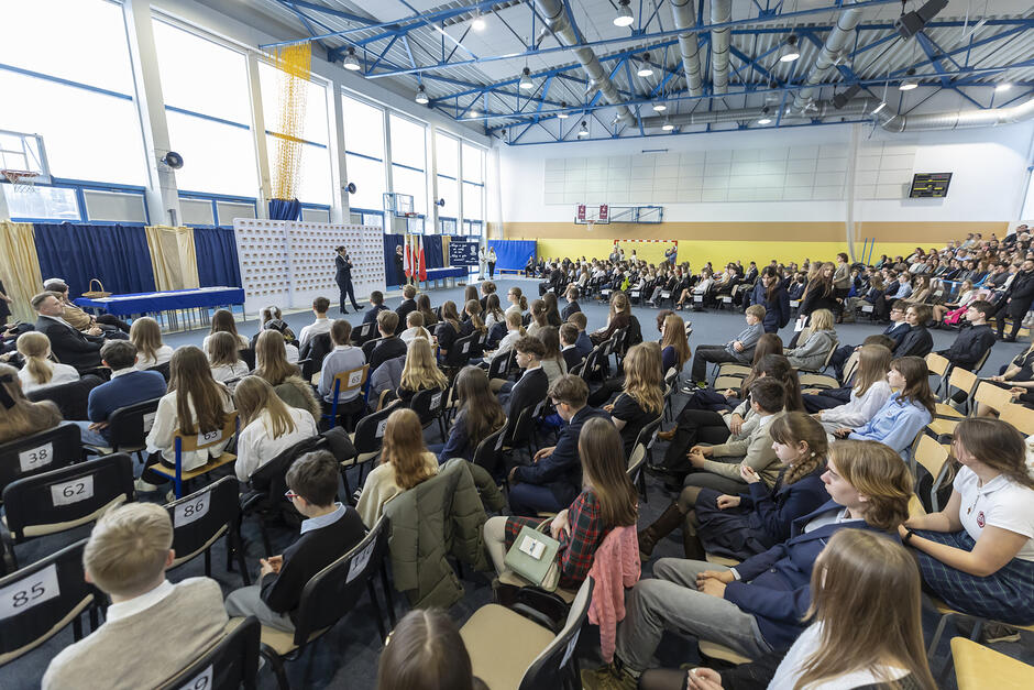 Na sali gimnastycznej siedzą setki uczniów. Przed nimi stoi kobieta i przemawia.