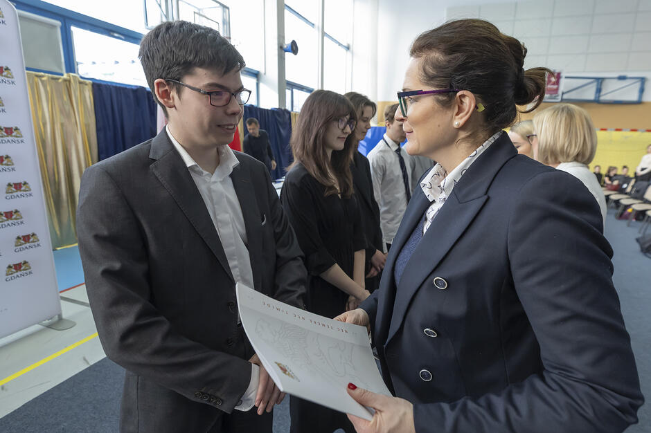 Kobieta wręcza stypendium uczniowi w marynarce na sali gimnastycznej w szkole.