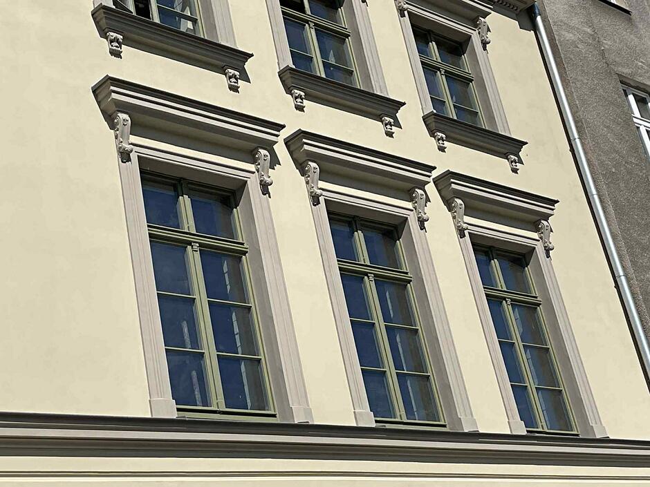 Na zdjęciu widać fragment jasnej elewacji zabytkowej kamienicy przy ulicy Łąkowej 28 w Gdańsku. Charakterystycznym elementem są trzy wysokie okna z zieloną stolarką, ozdobione klasycznymi gzymsami i dekoracyjnymi