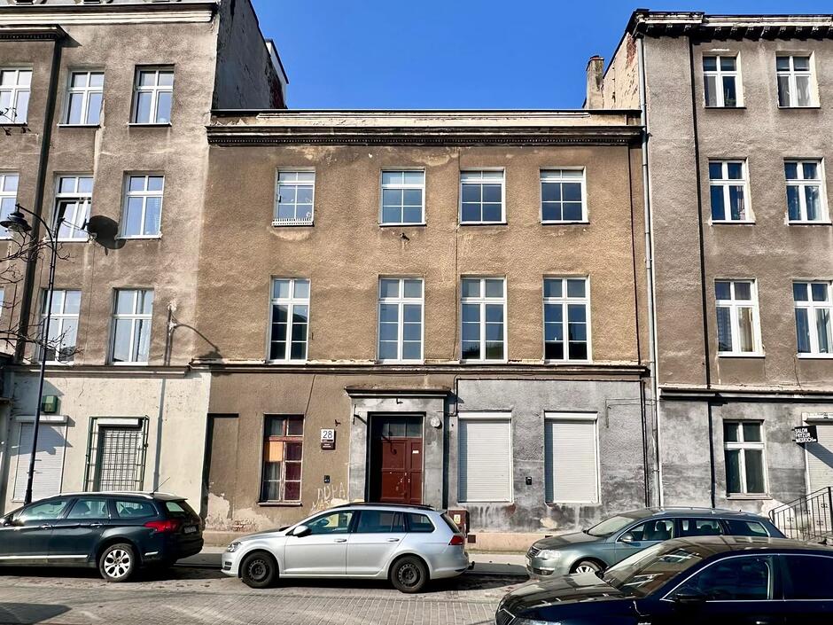 Zdjęcie przedstawia trzypiętrową kamienicę o surowej, tynkowanej elewacji z widocznym numerem 28, znajdującą się przy ulicy Łąkowej w Gdańsku. Budynek posiada prostą formę z regularnie rozmieszczonymi oknami i jest otoczony przez parkujące na chodniku samochody oraz sąsiednie, wyższe kamienice.
