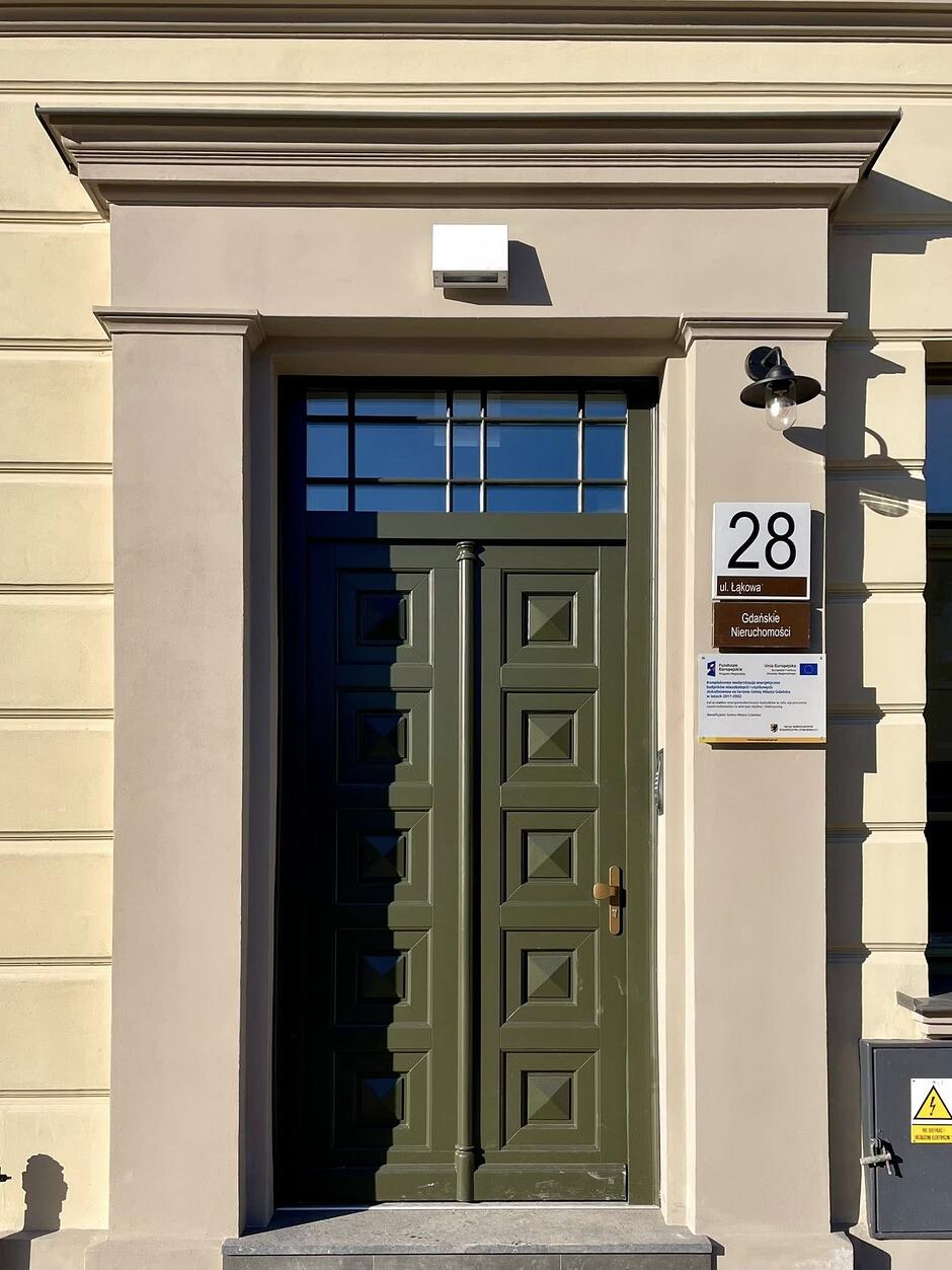 Zdjęcie przedstawia wejście do odrestaurowanej kamienicy przy ulicy Łąkowej 28 w Gdańsku, które wyróżnia się eleganckimi, ciemnozielonymi drzwiami z ozdobnymi kasetonami. Nad drzwiami znajduje się przeszkolone nadproże, a obok umieszczono tabliczki informacyjne zarządcy Gdańskie Nieruchomości oraz funduszy unijnych.