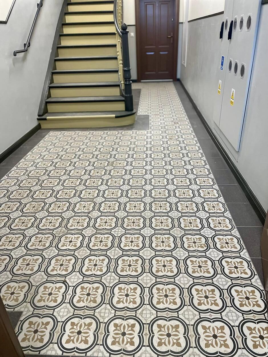 Zdjęcie przedstawia odnowione wnętrze klatki schodowej w zabytkowej kamienicy przy ulicy Łąkowej 28 w Gdańsku. Charakterystycznym elementem korytarza jest podłoga wyłożona dekoracyjnymi płytkami gresowymi o patchworkowym wzorze w stylu retro, które współgrają z klasycznymi, drewnianymi schodami.