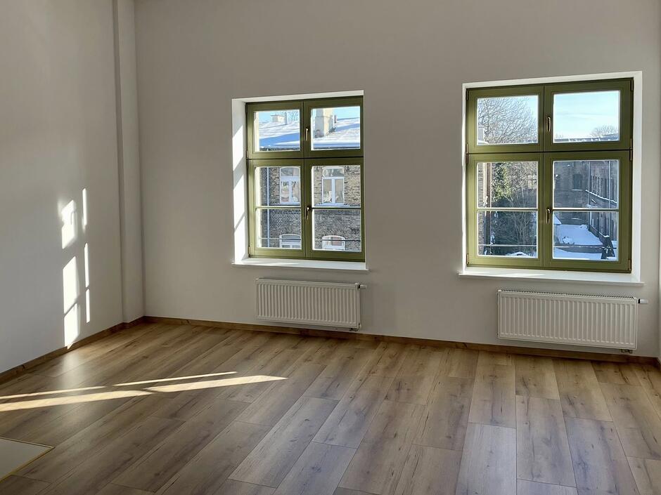 Zdjęcie przedstawia puste, nowo wyremontowane wnętrze pokoju w kamienicy przy ulicy Łąkowej 28 w Gdańsku. W jasnym pomieszczeniu znajdują się dwa duże okna o charakterystycznych zielonych ramach, pod którymi zamontowano nowoczesne grzejniki, a na podłodze ułożono panele o wzorze jasnego drewna.
