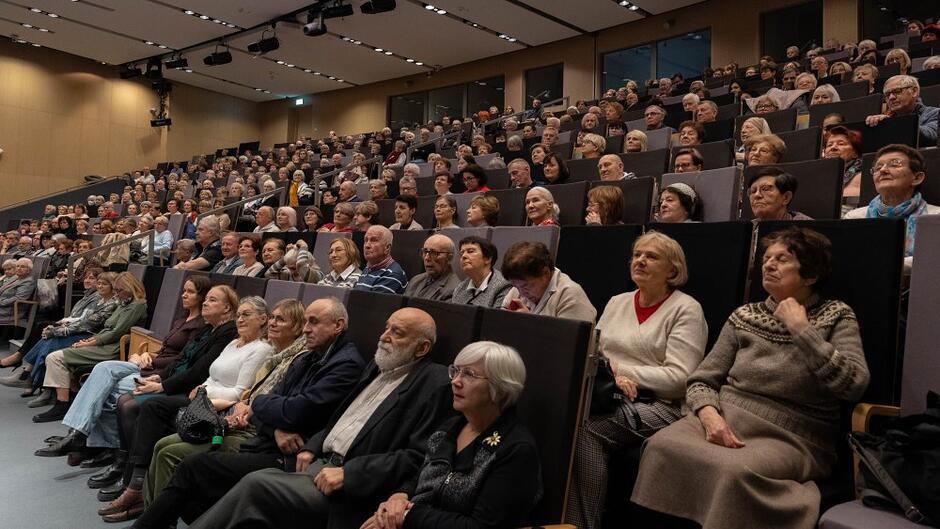 Grupa kilkuset seniorów siedzi na sali wykładowej i patrzy w jedną stronę. Prawdopodobnie są na zajęciach