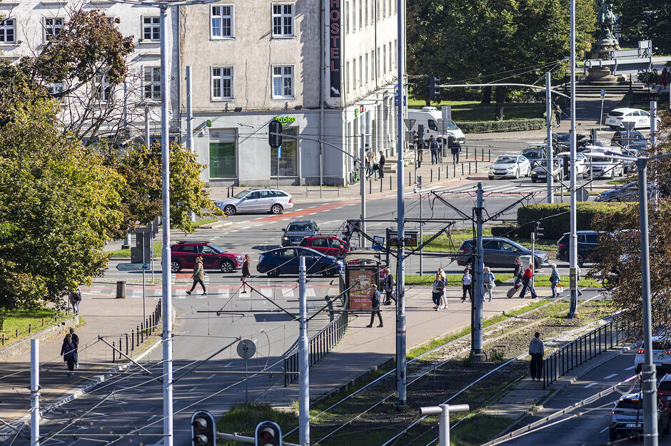 Zdjęcie przedstawia ruchliwe skrzyżowanie ulic , widoczne z perspektywy lotu ptaka w słoneczny dzień. Na pierwszym planie widać rozbudowaną infrastrukturę tramwajową z gęstą siecią trakcyjną oraz pieszych korzystających z przejść w pobliżu budynków użyteczności
