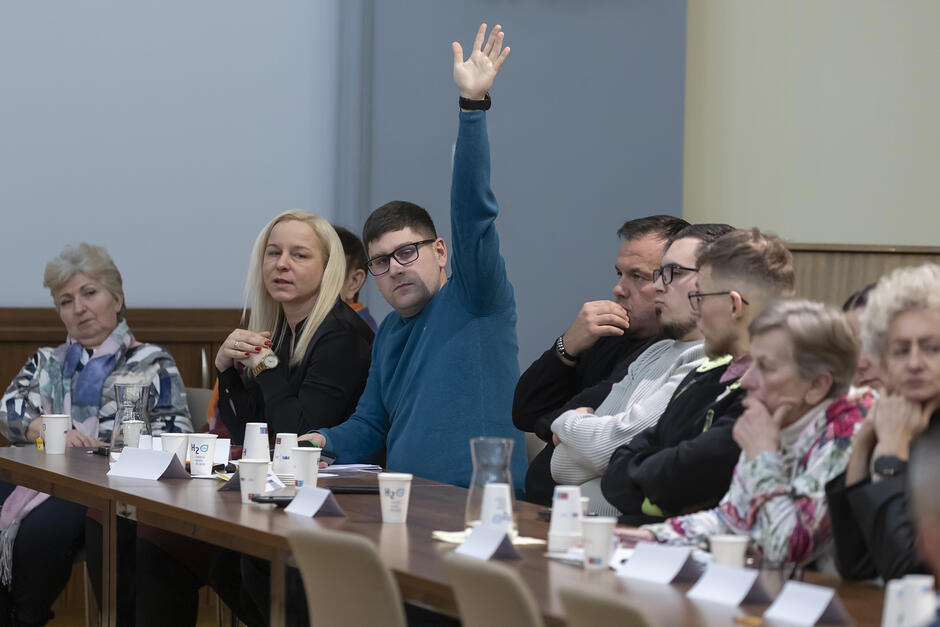 kilka osób siedzących przy stole w trakcie debaty. Jedna z nich - mężczyzna - podnosi rękę zgłaszając gotowość do zabrania głosu 