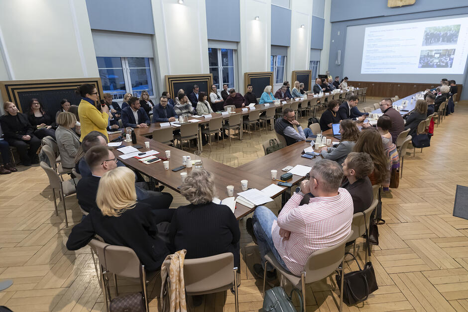 Wielki stół w obszernej sali. Stół składa się z mniejszych stołów ustawionych w literę U. Przy stołach siedzą uczestnicy spotkania 