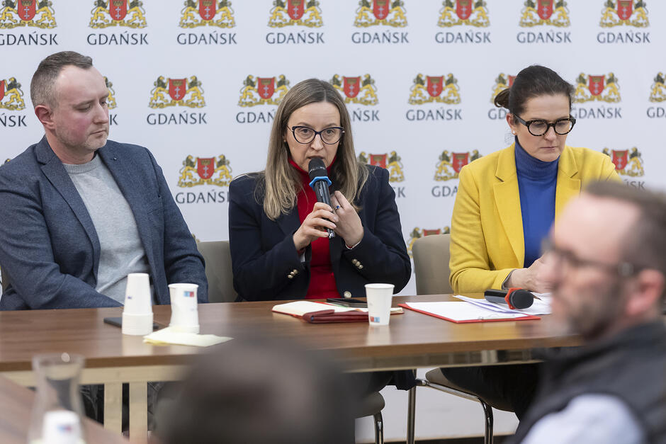 Trzy osoby siedzące przy stole. Pośrodku przemawia kobieta z mikrofonem w ręku 