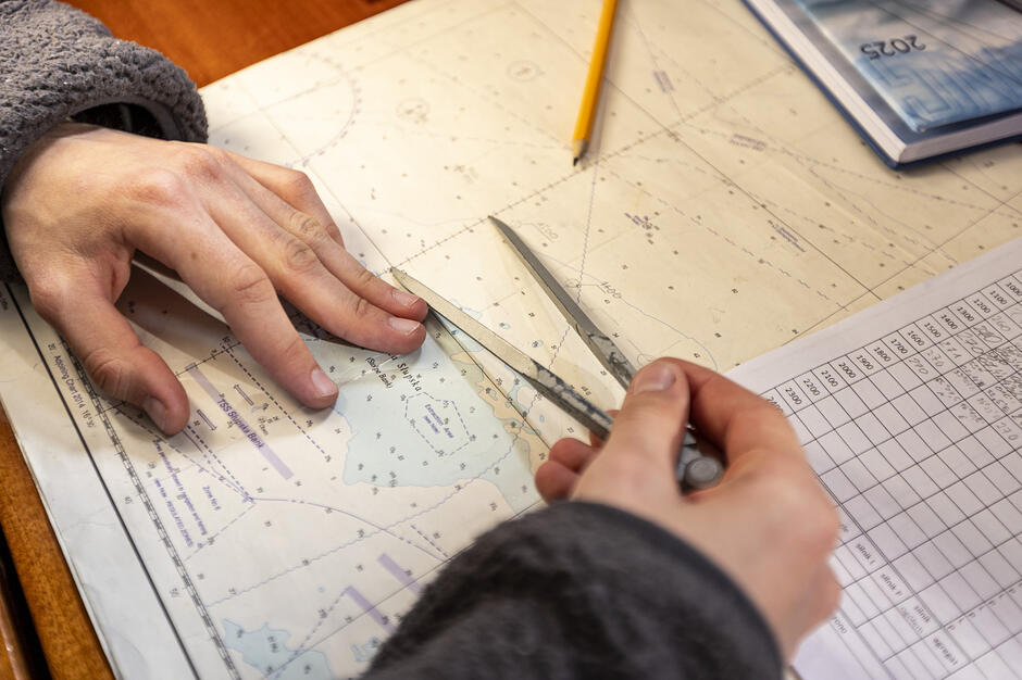 Zdjęcie przedstawia ręce osoby pracującą z mapą nawigacyjną, na której odmierza odległości za pomocą cyrkla traserskiego. W tle widoczne są przybory biurowe, takie jak ołówek i notatki z tabelami, co sugeruje planowanie kursu lub naukę nawigacji.