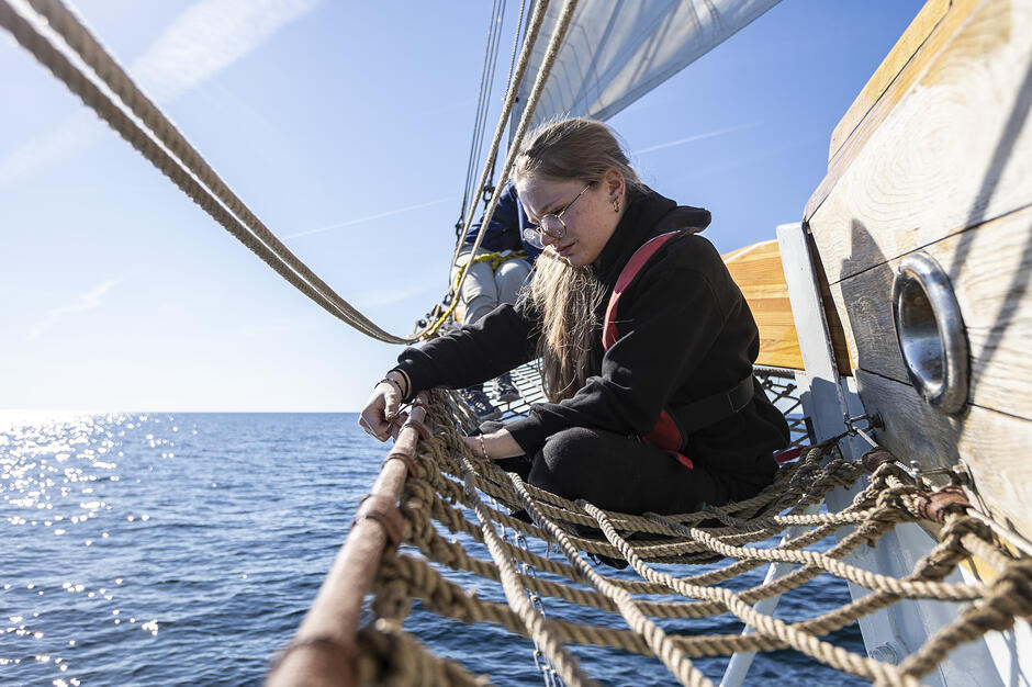 Zdjęcie przedstawia młodą kobietę siedzącą w siatce pod bukszprytem żaglowca, która zajmuje się pracami bosmańskimi przy linach. W tle rozpościera się widok na spokojne, błękitne morze w pełnym słońcu, co podkreśla żeglarski charakter fotografii.