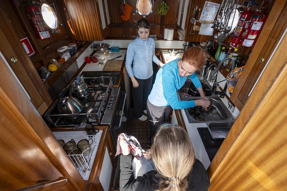 Zdjęcie przedstawia trzy kobiety pracujące w ciasnym kambuzie, czyli kuchni na jachcie lub żaglowcu. Widać, jak wspólnie zajmują się myciem i wycieraniem naczyń w otoczeniu profesjonalnego wyposażenia morskiego, takiego jak kuchenka z relingami i zawieszone naczynia.
