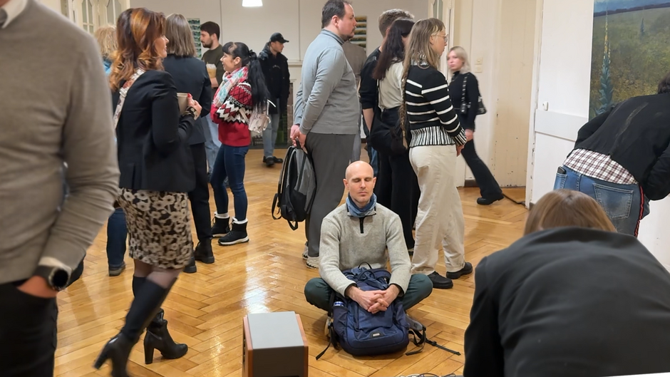 Zdjęcie przedstawia grupę ludzi w przestronnym pomieszczeniu z drewnianym parkietem, prawdopodobnie podczas jakiegoś wydarzenia lub spotkania. W centrum uwagi znajduje się mężczyzna siedzący na podłodze w pozycji medytacyjnej z zamkniętymi oczami, podczas gdy wokół niego stoją i rozmawiają inne osoby.