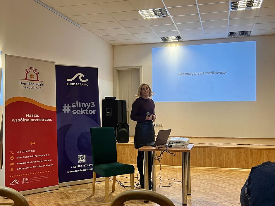 Zdjęcie przedstawia prelekcję w Domu Sąsiedzkim „Zakopianka” dotyczącą podstaw prawa spadkowego. W centrum widać kobietę prowadzącą wykład przy stoliku z laptopem i rzutnikiem, na tle banerów informacyjnych Fundacji RC.