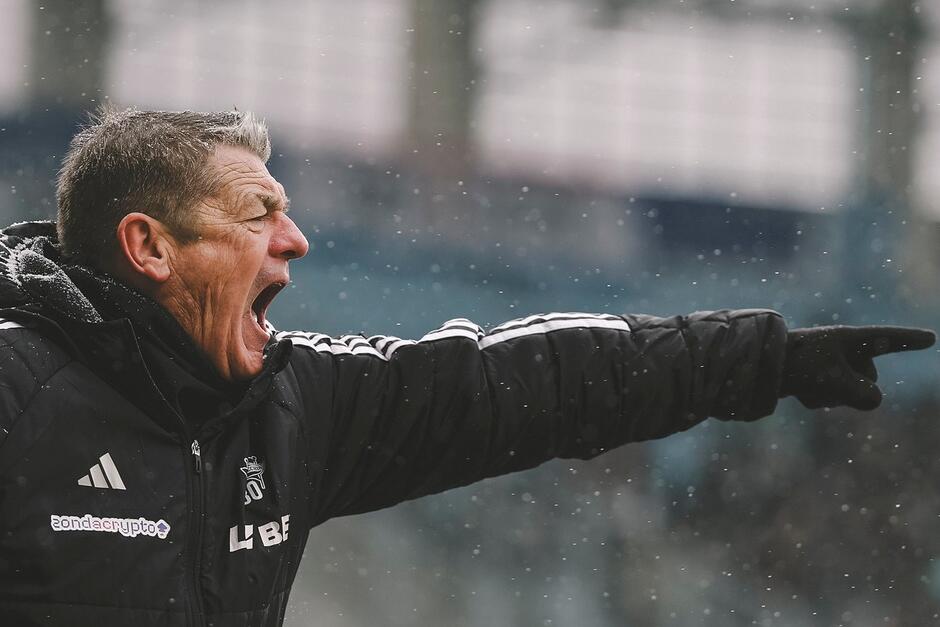 Mężczyzna lat około 60 krzyczy stojąc przy linii bocznej boiska podczas meczu piłki nożnej. Jest ubrany w ciepłą czarną kurtkę 