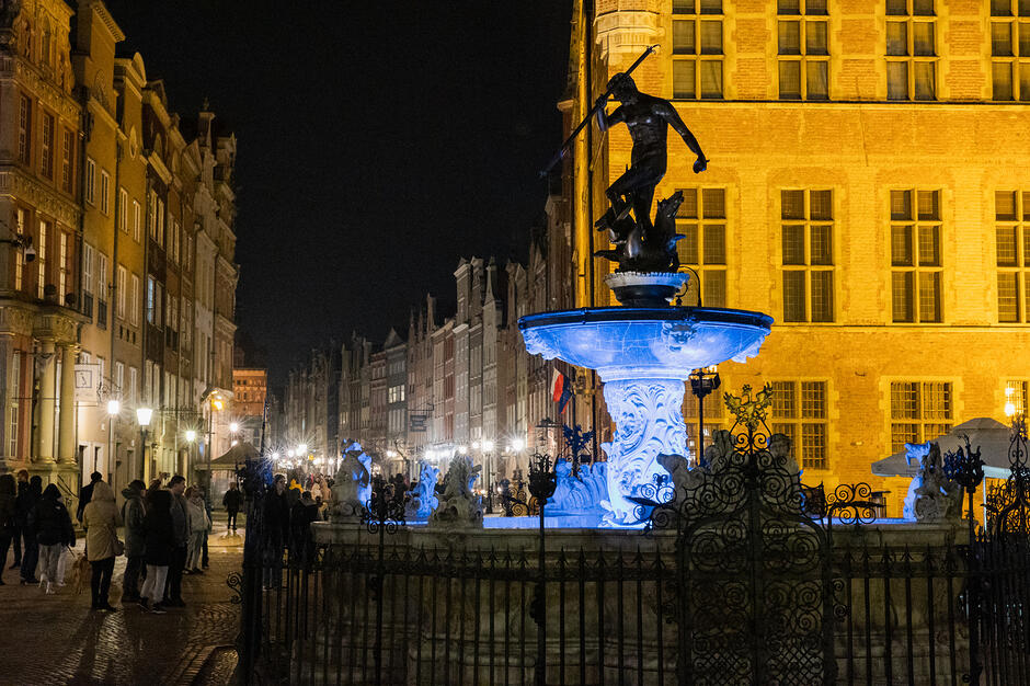zdjęcie zrobione po zmroku, widać zabytkową fontannę neptuna podświetloną na niebiesko, w tle zabytkowe oświetlone kamienice