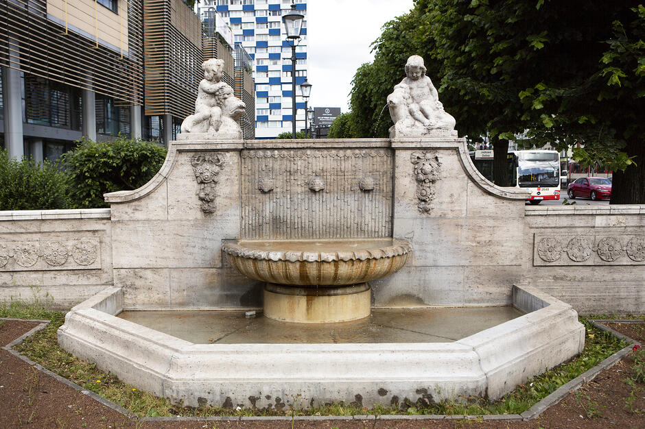 na zdjęciu jasna niewysoka fontanna, w tle po lewej i z tyłu budynki