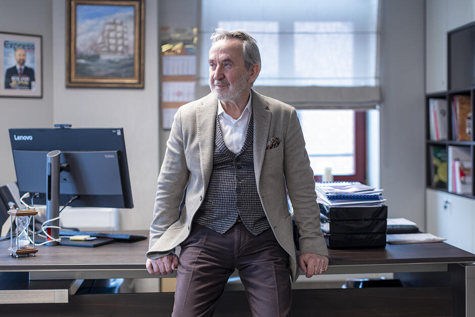 elegancko ubrany mężczyzna w podeszłym wieku, opiera się o biurko na którym widać ekran komputera i stos dokumentów