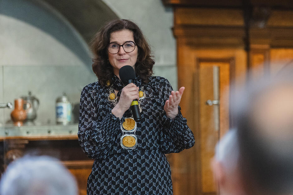 elegancko ubrana kobieta w okularach i niebieskiej sukience. Ma w dłoniach mikrofon, mówi, prawdopodobnie do osób zgromadzonych