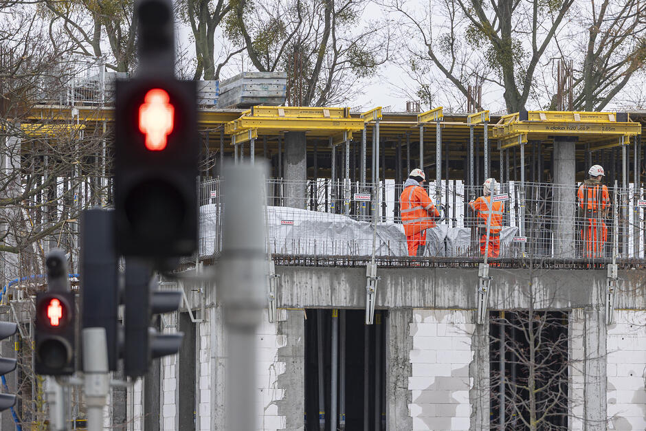 robotnicy w pomarańczowych kombinezonach stoją na budowanym betonowym budynku