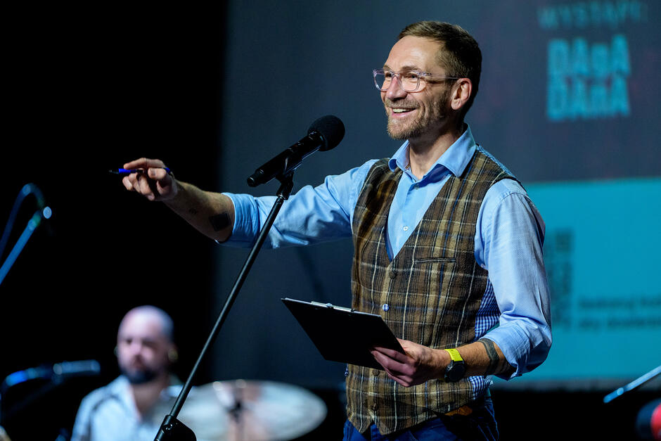 Na zdjęciu widać uśmiechniętego mężczyznę w okularach, ubranego w błękitną koszulę i kraciastą kamizelkę, który występuje na scenie z mikrofonem. W jednej ręce trzyma podkładkę z dokumentami i długopis, a drugą gestykuluje w stronę publiczności, podczas gdy w tle widoczny jest perkusista.