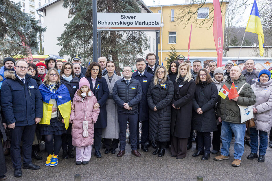 Liczna grupa osób pozuje do fotografii na Skwerze Bohaterskiego Mariupola, gdzie zapalono znicze. Widać ukraińskie flagi.