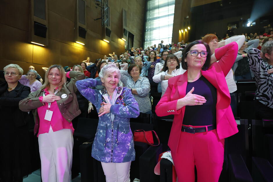 kobiety stoją na dużym audutorium, są w różnym wieku, wykonują samobadanie piersi