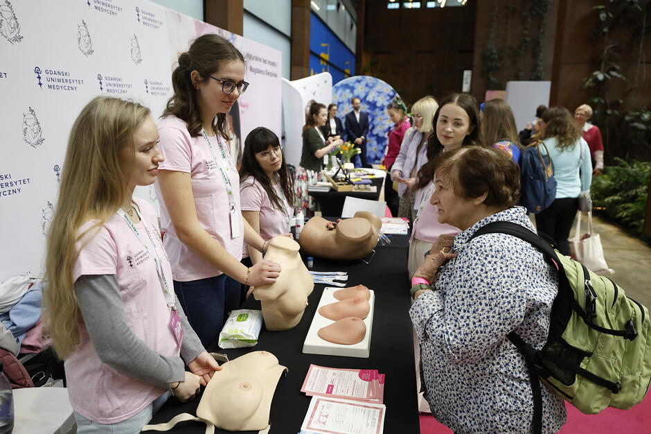 Na zdjęciu widać stoisko Gdańskiego Uniwersytetu Medycznego podczas Gdańskiego Kongresu Zdrowia Kobiet, gdzie studentki położnictwa edukują uczestniczkę w zakresie profilaktyki raka piersi. Na pierwszym planie znajdują się fantomowe modele piersi służące do nauki prawidłowej techniki samobadania