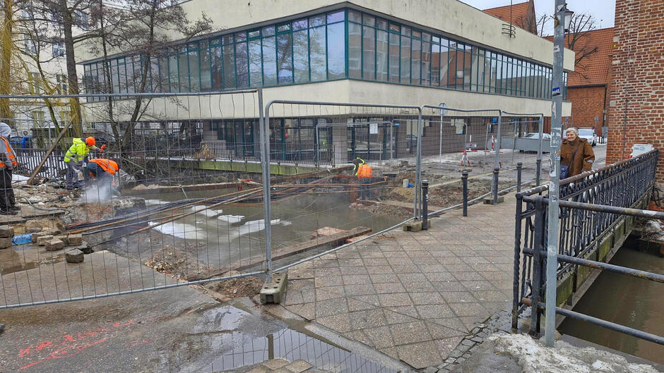 na zdjęciu ogrodzenie z siatki i chodnik, wokół kamienice