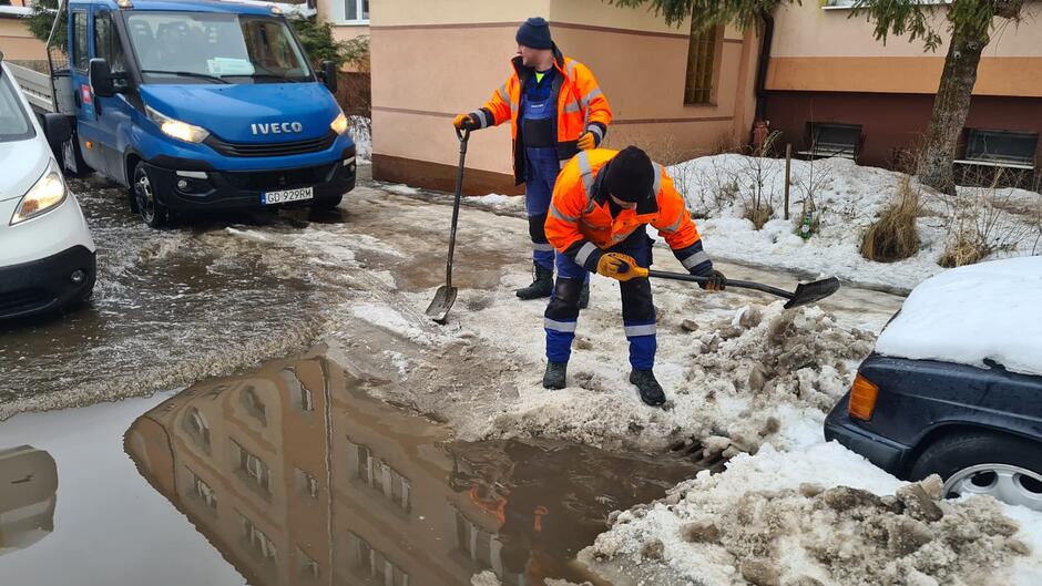 na zdjęciu dwóch mężczyzn z łopatami, jeden odśnieża ulicę, widać stojące obok nich auta