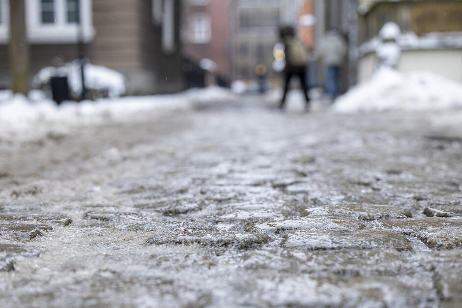 ulica pokryta lodem. W tle widoczne są domy i budynki. Na pierwszym planie znajduje się osoba chodząca po ulicy, a ziemia jest pokryta warstwą lodu po obu stronach drogi