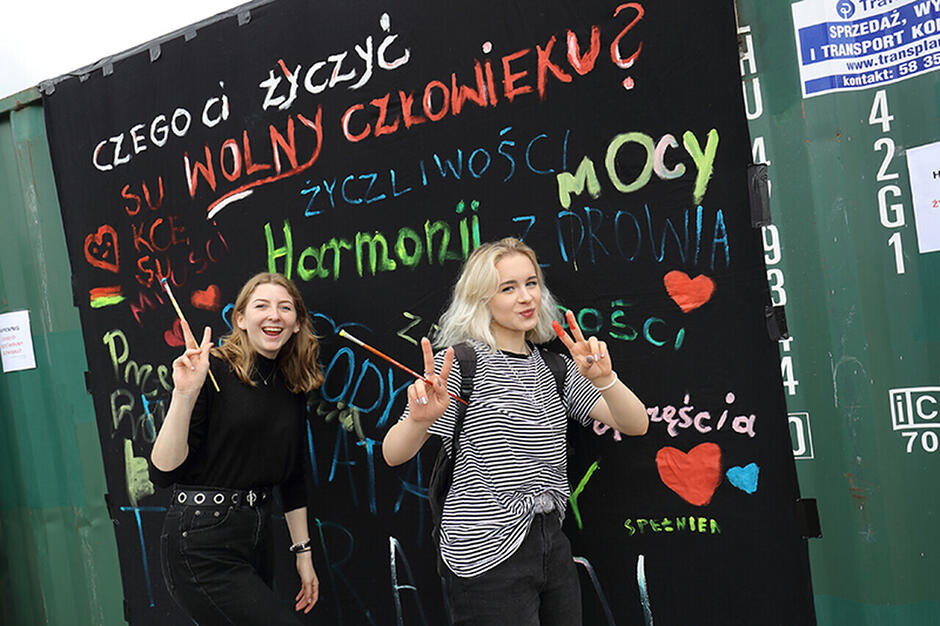 Dwie kobiety pozują do zdjęcia, w tle czarna tablica, na której napisane są różne hasła